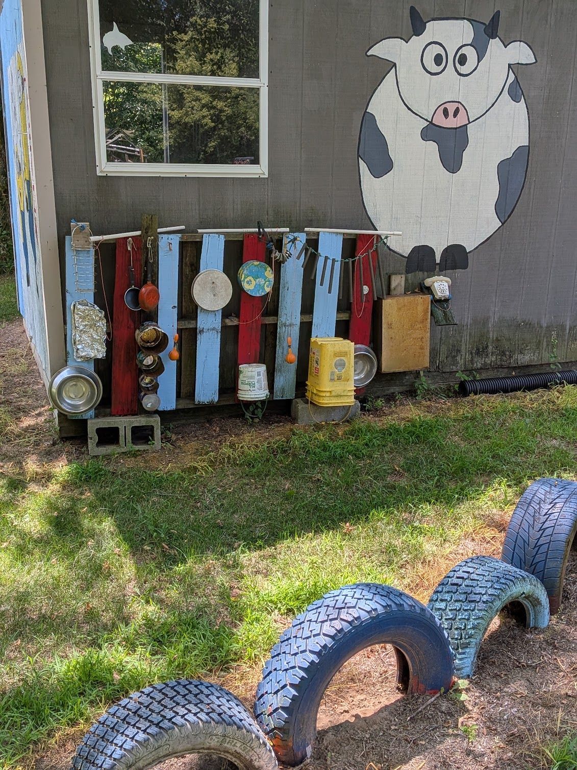 Painted cow mural beside a backyard fence with colorful reclaimed wooden planks and tire arches on grass