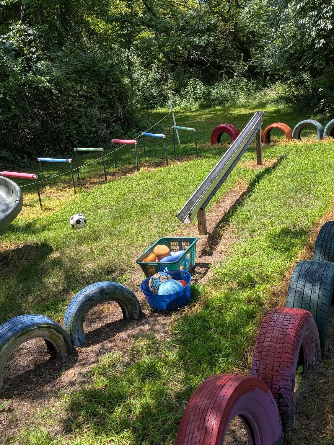 Playground obstacle course with tires, ramps, and a tunnel slide in a grassy, wooded area