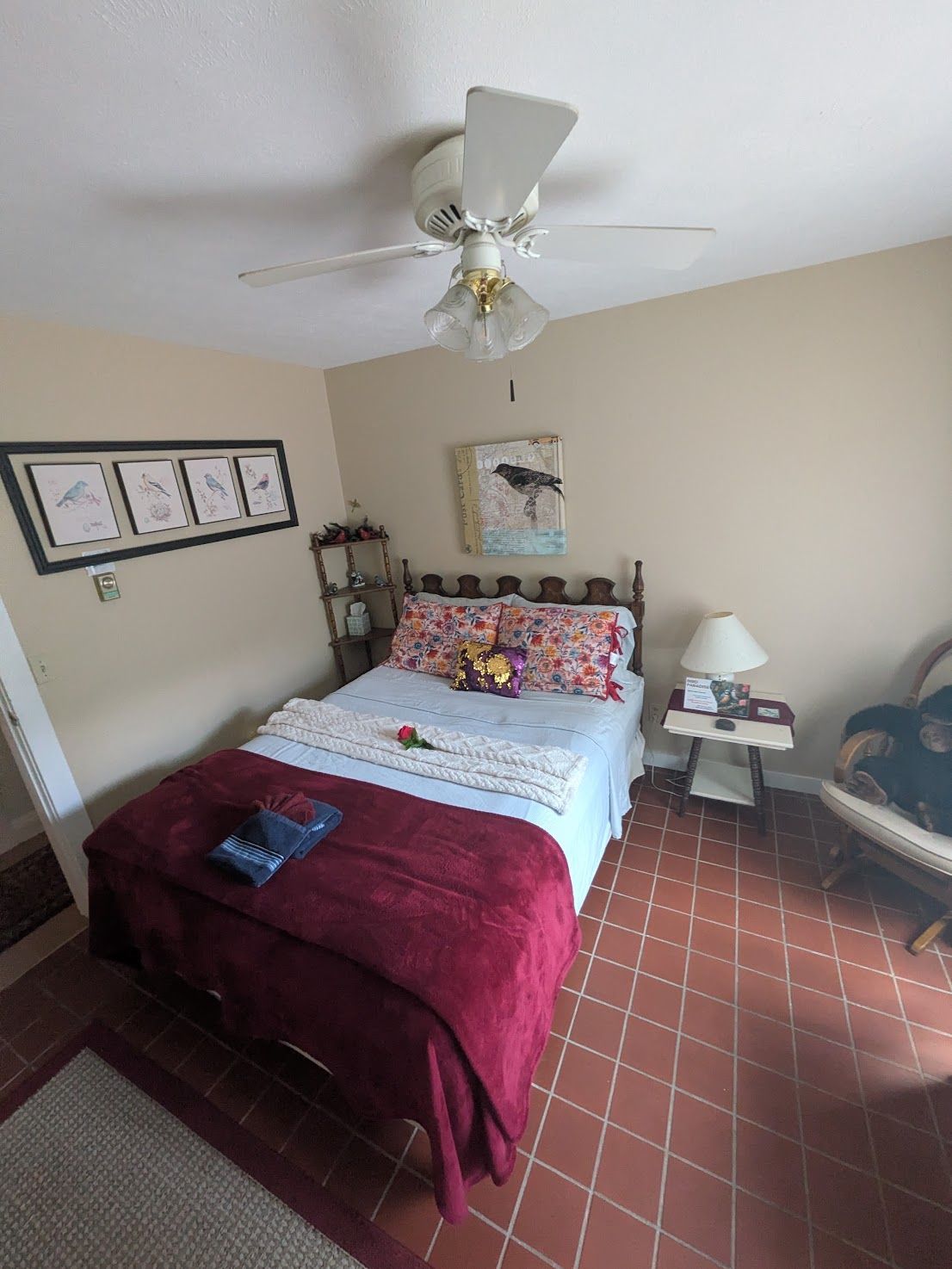 Bedroom with white bed, burgundy blanket, patterned pillows, ceiling fan, and beige walls.