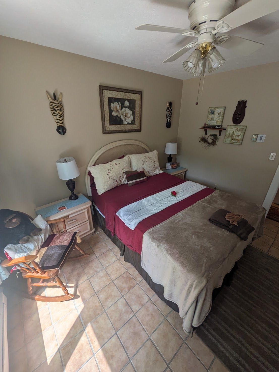 Small bedroom with a bed, ceiling fan, tiled floor, bedside table, and wall decor.