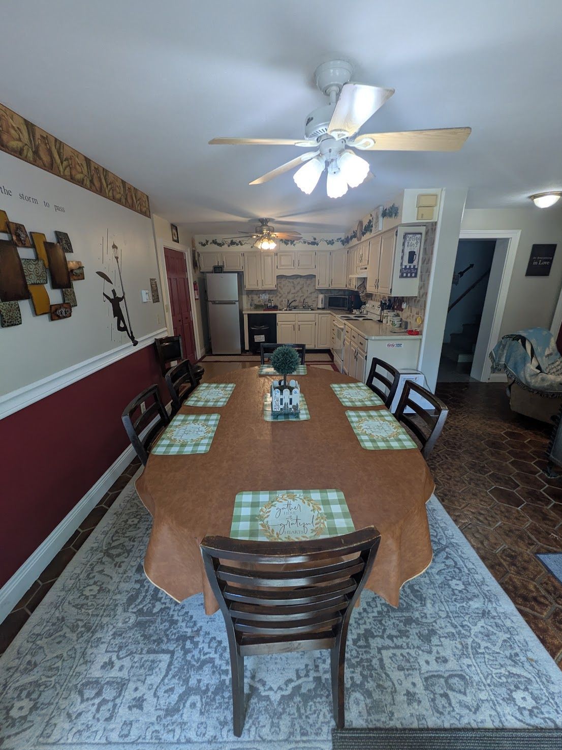 Dining room with a long table, chairs, ceiling fan, and kitchen visible in the background.