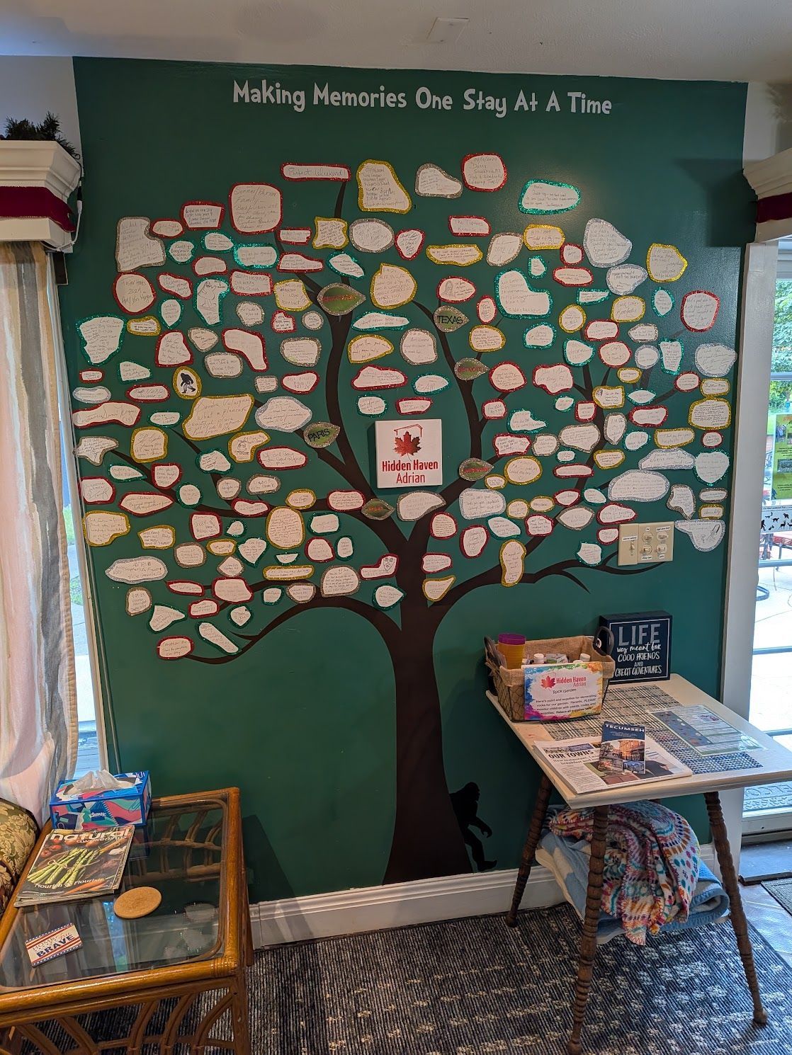 Classroom wall mural of a tree covered in leaves with students' handprints and books on tables nearby