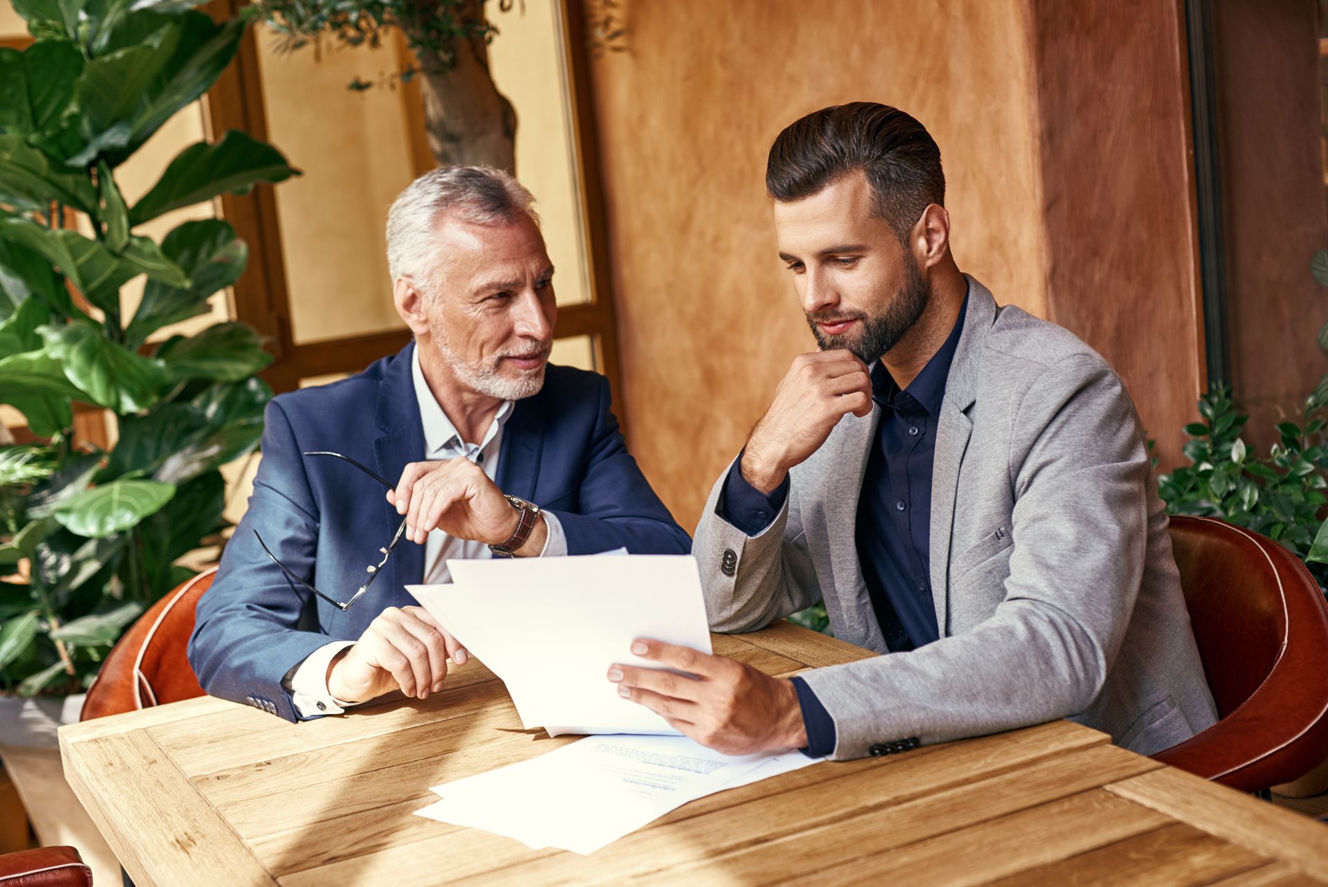 Two men in suits review a document at a wooden table in a warm office setting.