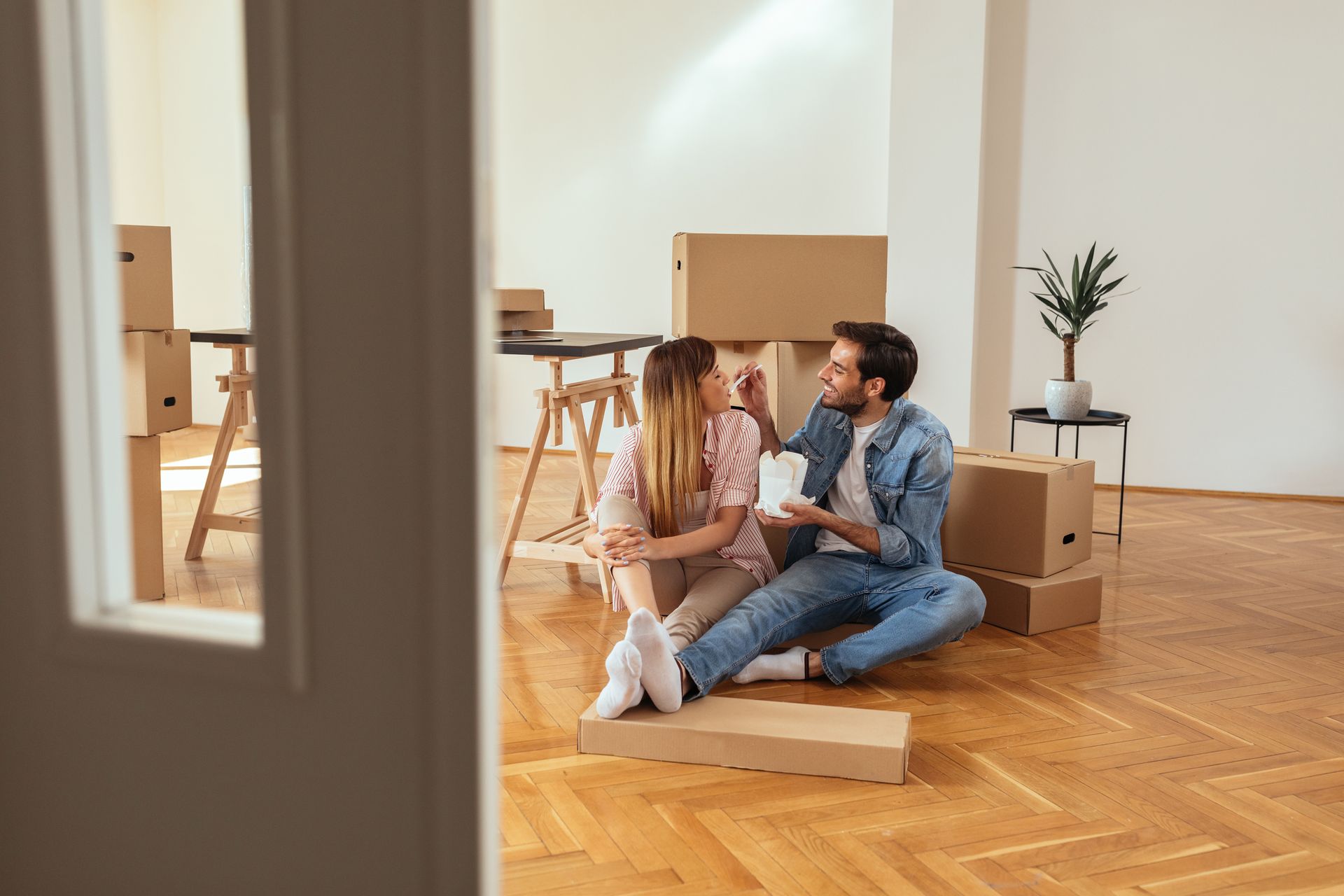 Couple sitting on the floor among moving boxes in a bright, empty room, smiling at each other