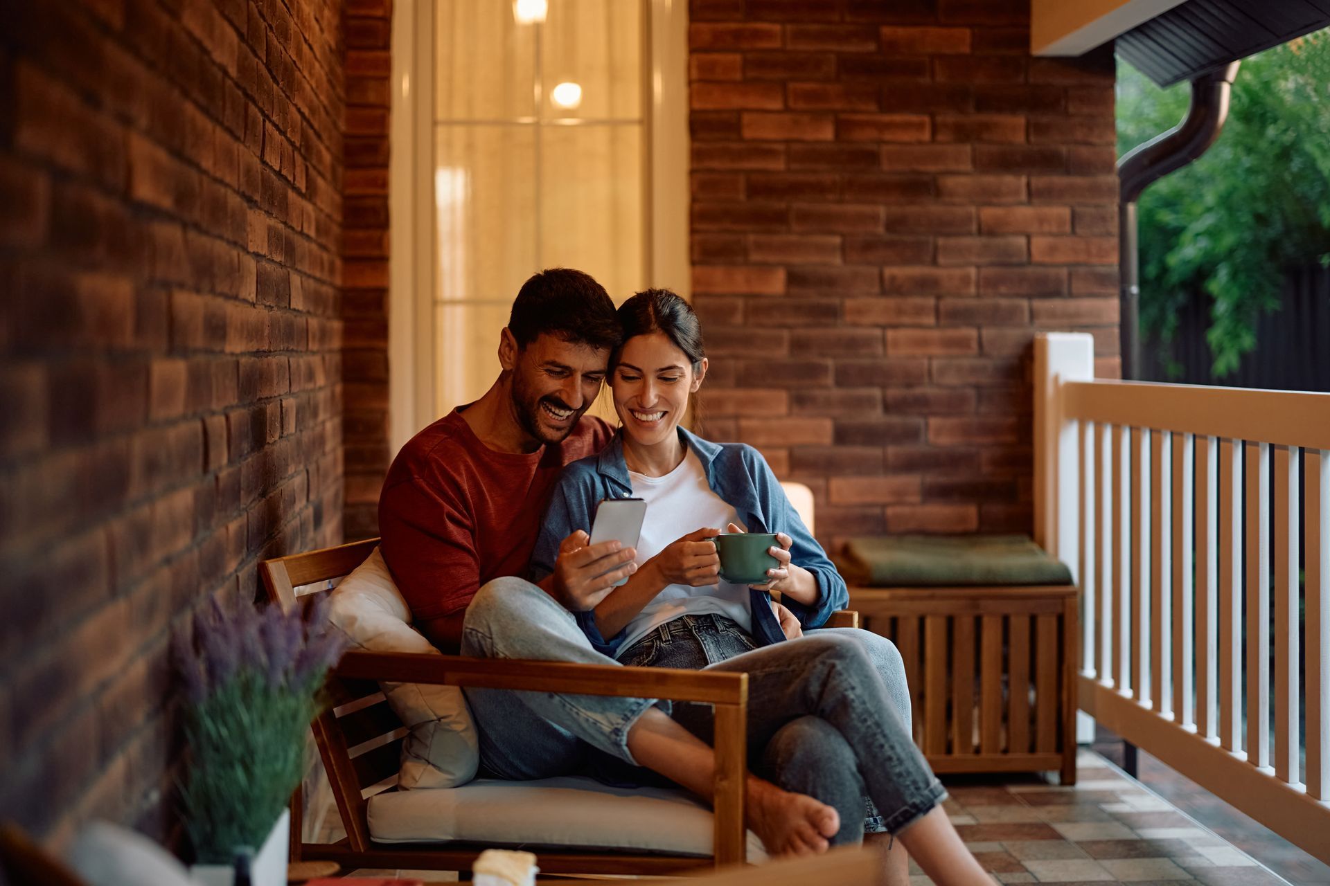 Couple relaxing on a porch, smiling at a tablet, with a baby on the woman’s lap
