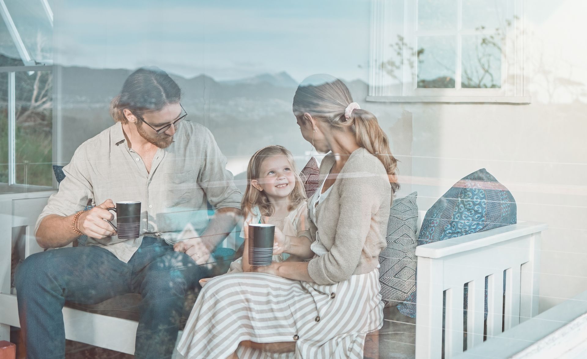 Family chatting on a bright porch, seated around a table with mugs and a child in the middle.