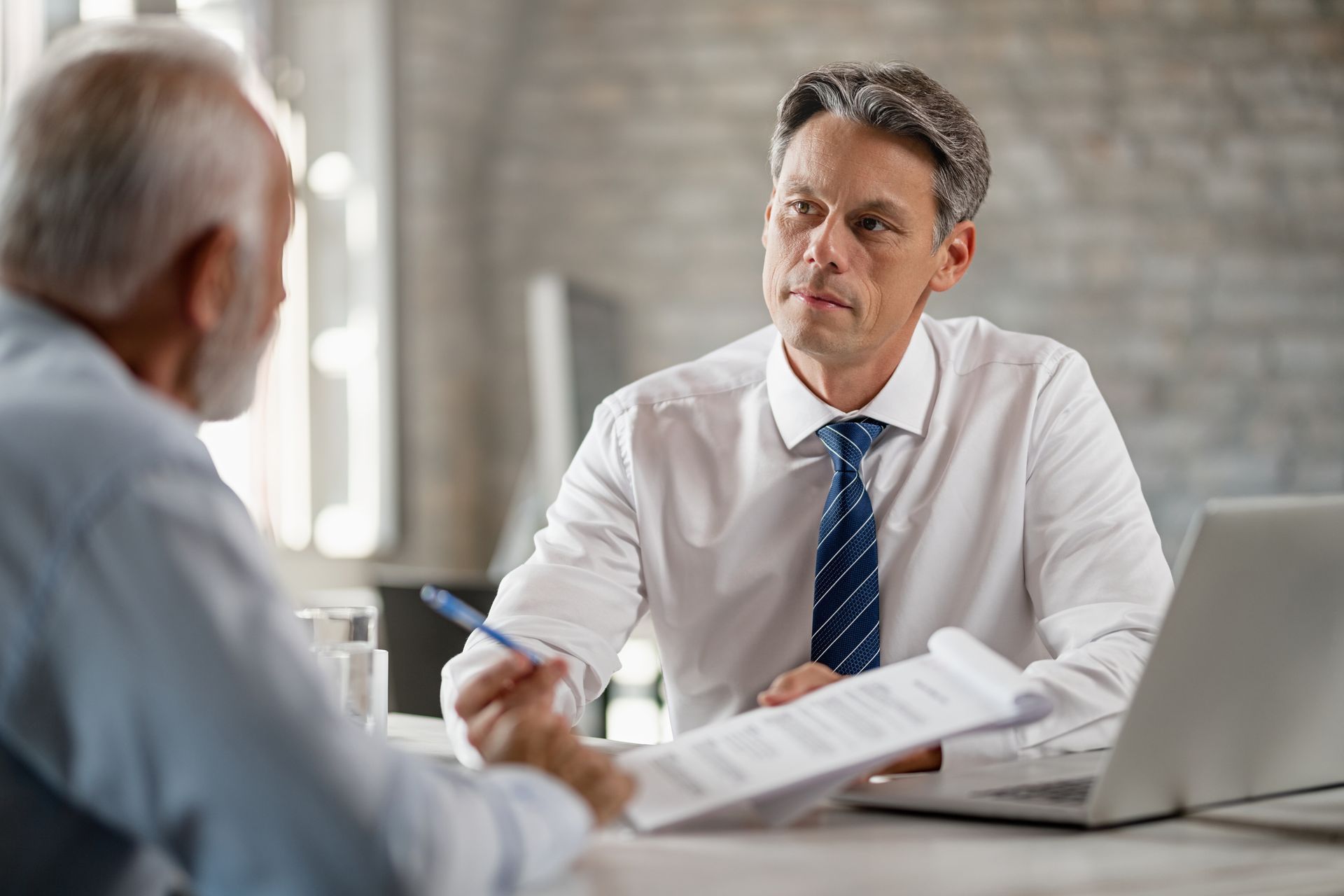 Two people in a business meeting, with one man speaking across a desk beside a laptop and documents.