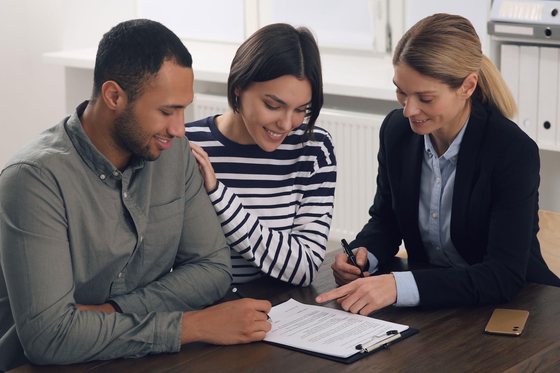 Three people review documents at a table, one pointing at a notebook in a bright office.