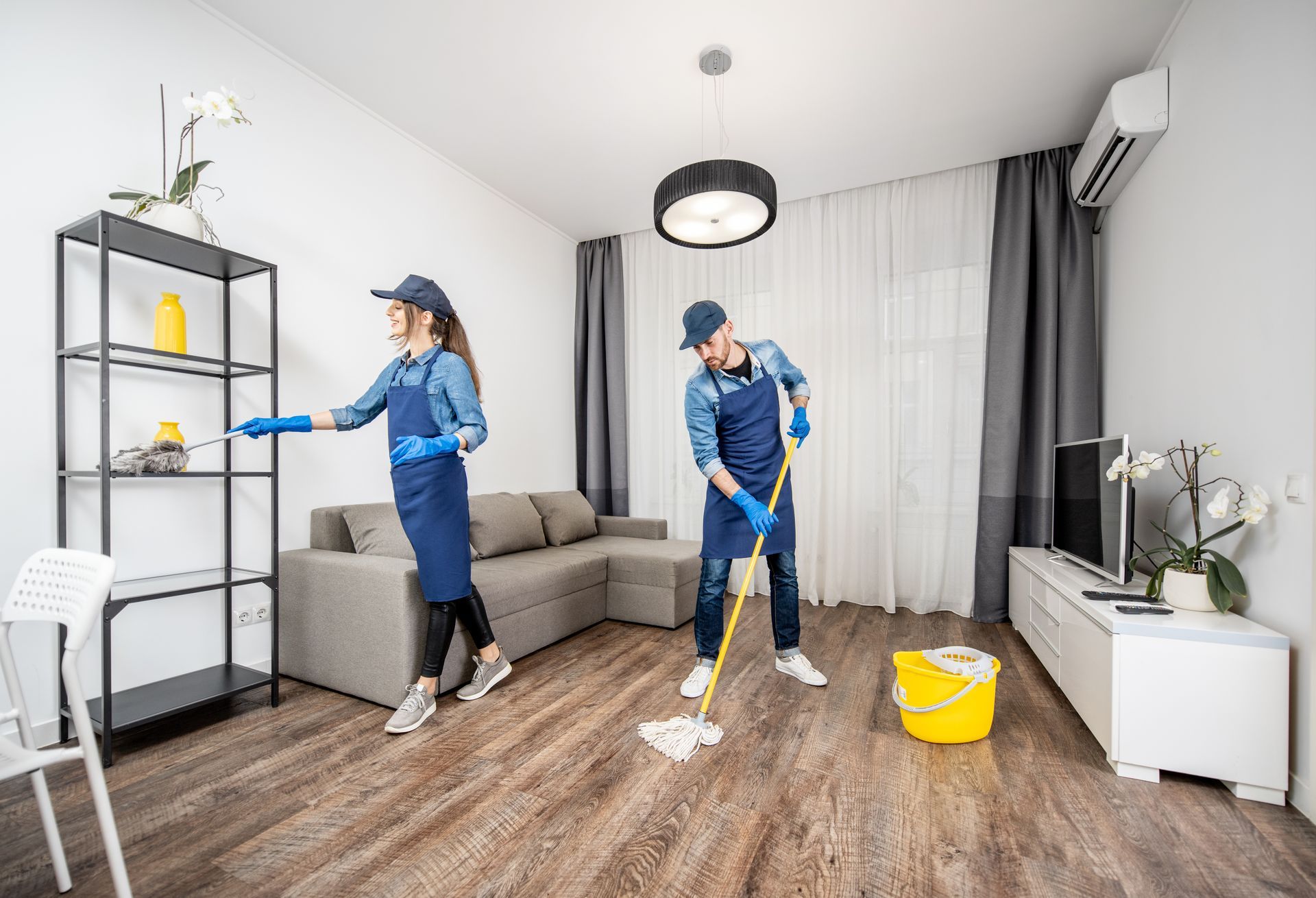 Two people in blue uniforms and gloves clean a living room: one wipes a tall metal shelf while the other mops the floor. Two people in blue uniforms and gloves clean a living room: one wipes a tall metal shelf while the other mops the floor.