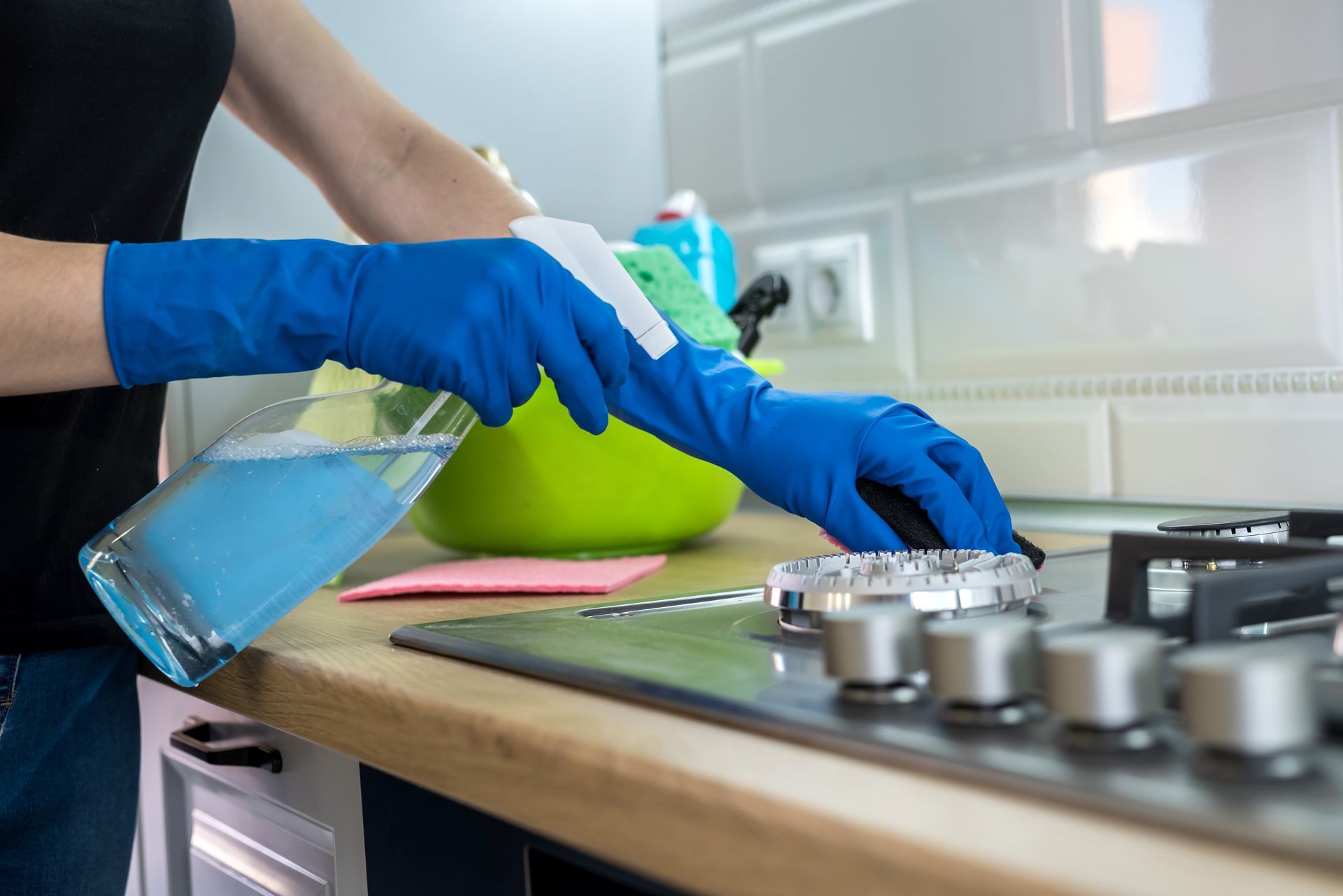 A person wearing blue rubber gloves uses a spray bottle and sponge to clean a stainless steel gas stovetop. A person wearing blue rubber gloves uses a spray bottle and sponge to clean a stainless steel gas stovetop.