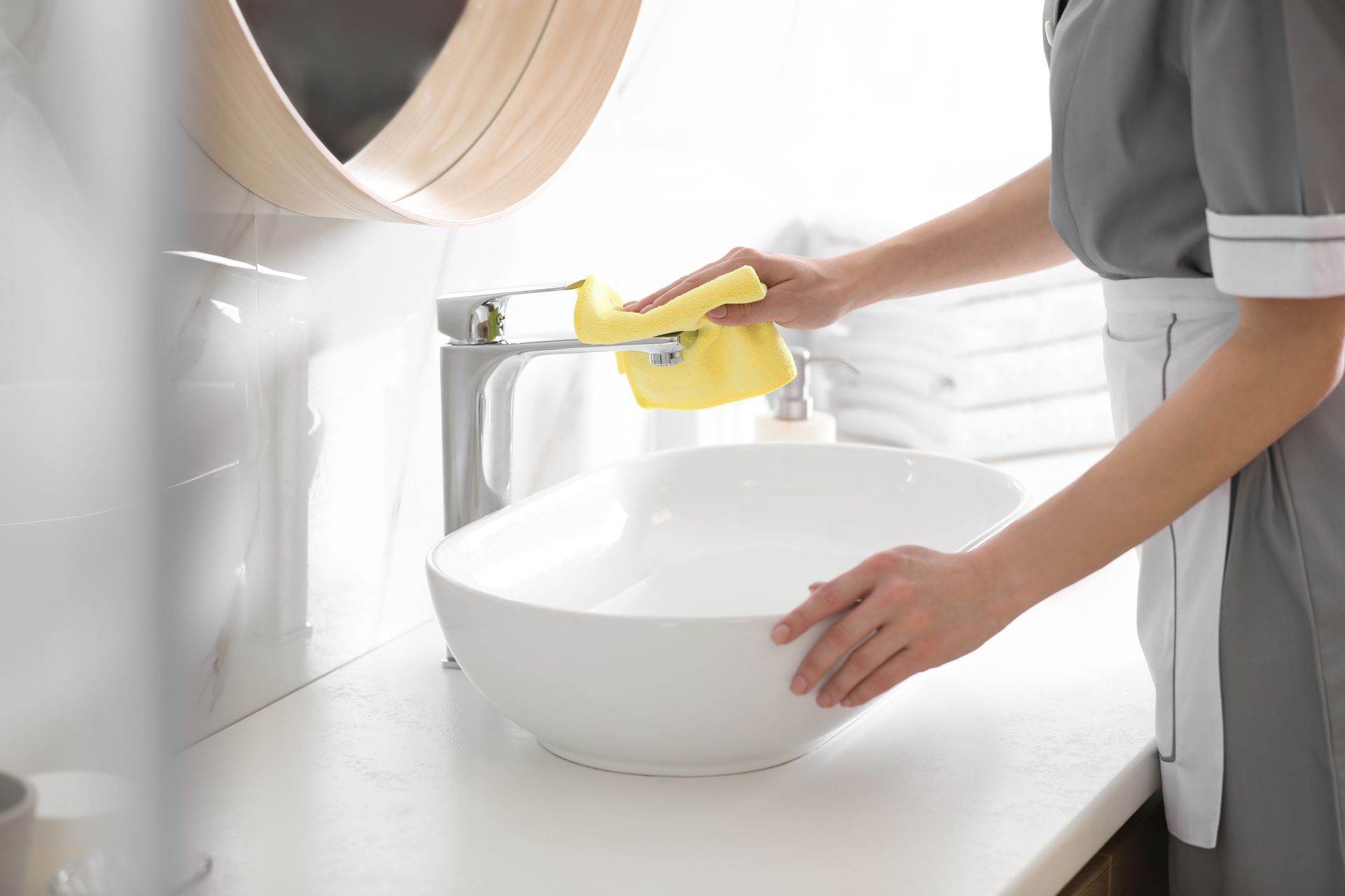 A person in a gray uniform cleans a chrome bathroom faucet with a yellow cloth above a white vessel sink. A person in a gray uniform cleans a chrome bathroom faucet with a yellow cloth above a white vessel sink.