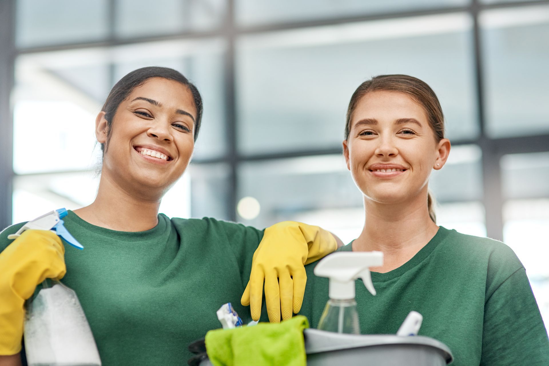 Two people wearing green shirts and yellow rubber gloves smile while holding cleaning supplies in an office setting. Two people wearing green shirts and yellow rubber gloves smile while holding cleaning supplies in an office setting.