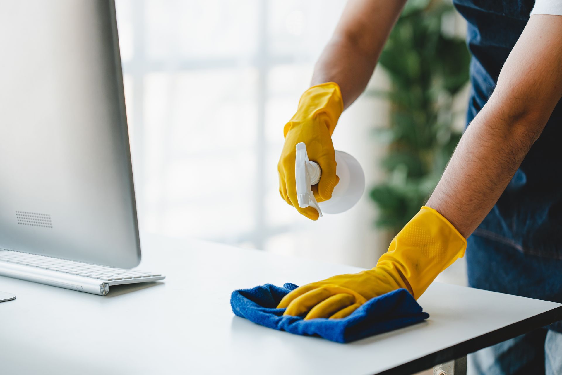 A person wearing yellow rubber gloves wipes a clean white desk with a blue cloth while holding a spray bottle.