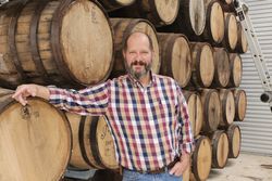 Man standing beside stacked wooden wine barrels, wearing a red and blue plaid shirt and jeans