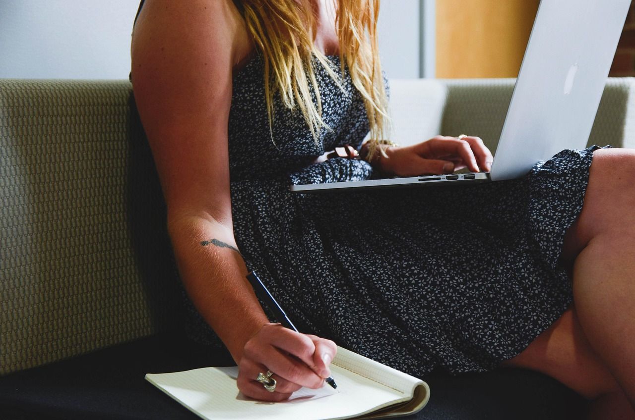 Woman in black dress typing on laptop, taking notes on a notepad while sitting on a couch.