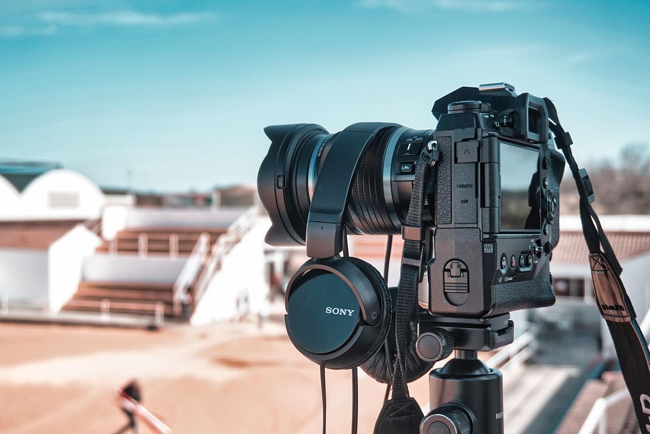 Camera on tripod with headphones; blurred background of a person and white structures under a blue sky.