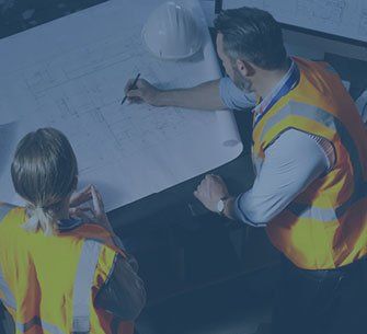 Two construction workers reviewing blueprints, one drawing with a pen, wearing safety vests.