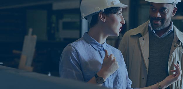 Two people in white hard hats discussing in a factory setting.