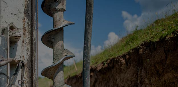 Close-up of a drilling auger against a blue sky, grass, and exposed earth.