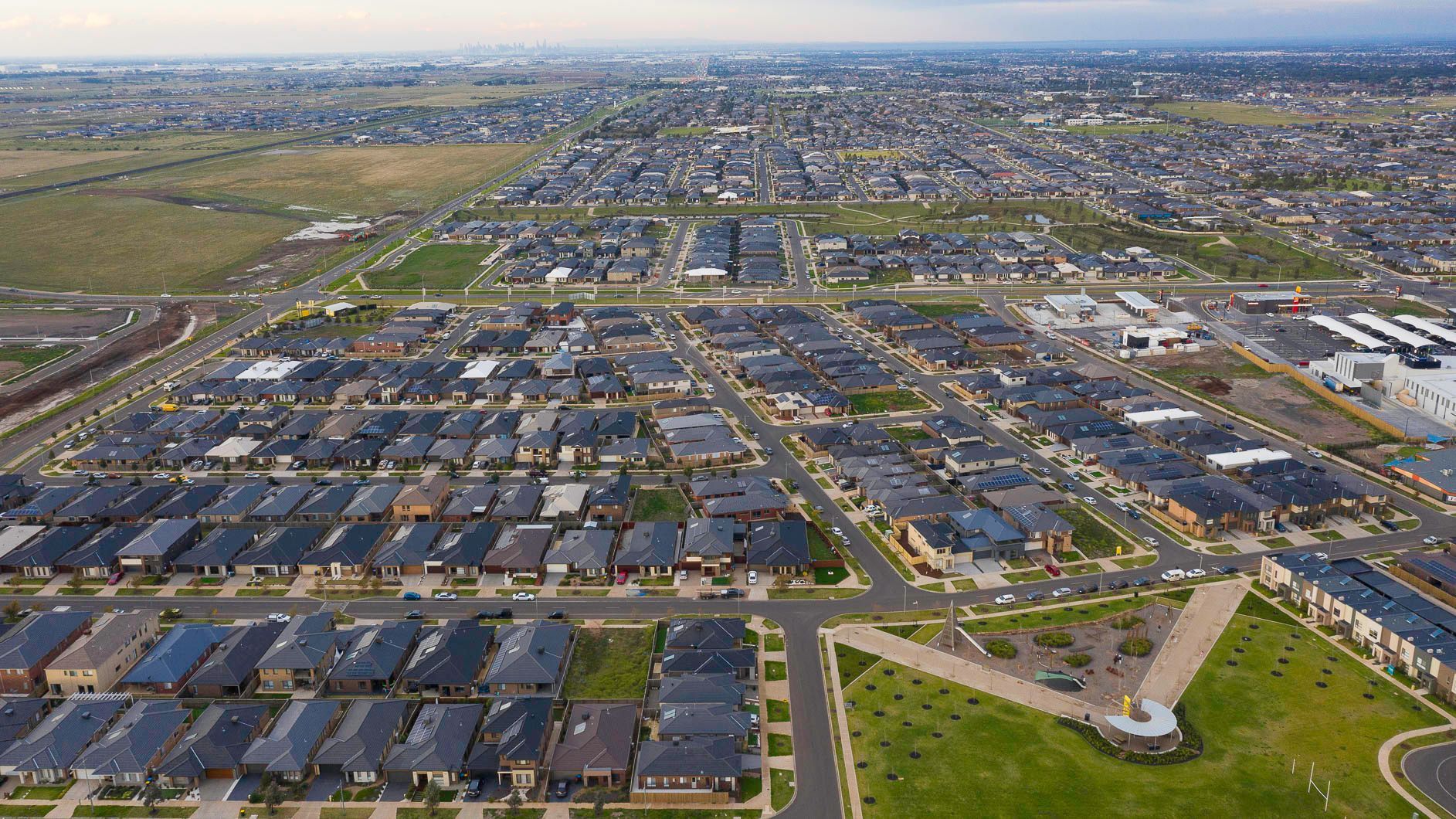 Aerial view of a suburban neighborhood with rows of houses, roads, and green spaces under an overcast sky.