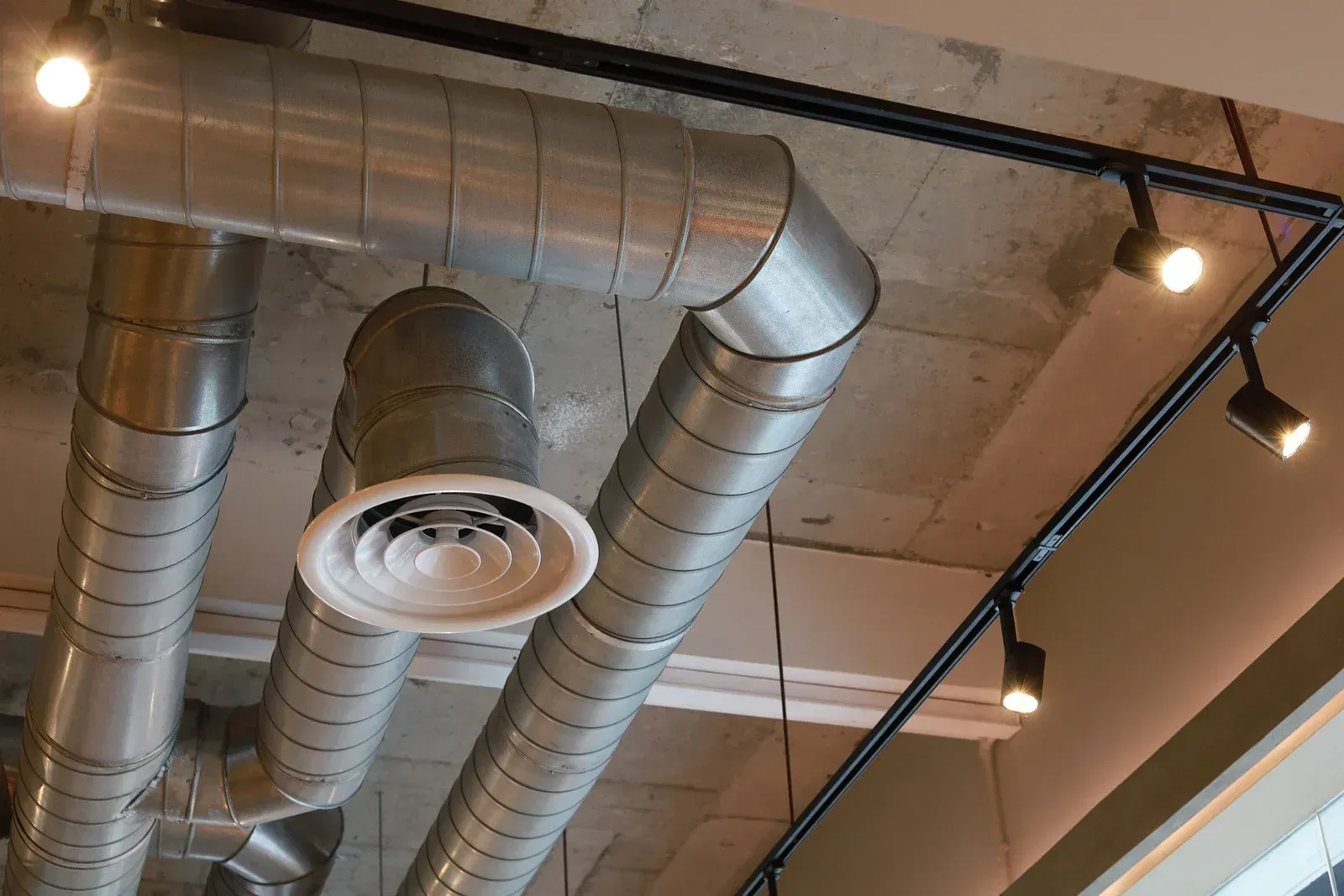 Silver HVAC ducts and black track lighting on a concrete ceiling in an industrial setting.