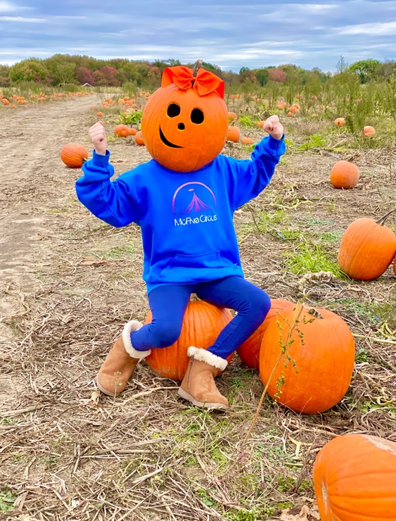 A person with a pumpkin head wearing a bright blue sweatshirt and tan boots sits on pumpkins in a field.