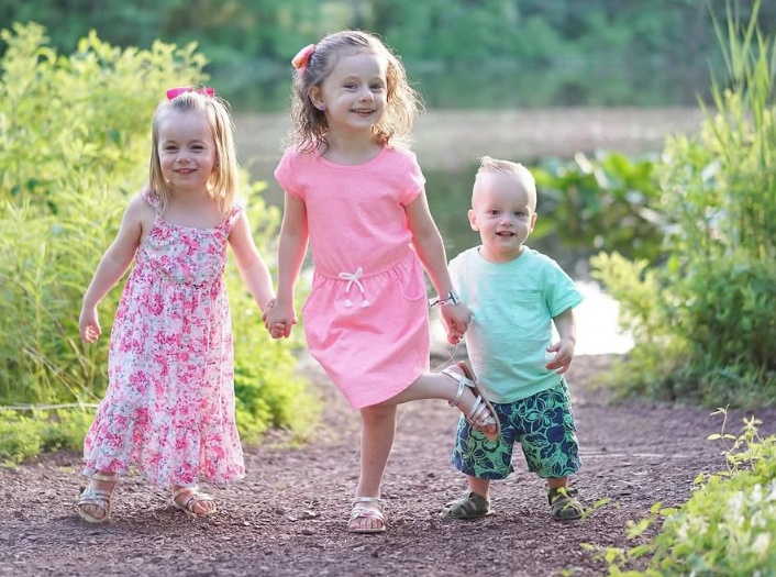 Two girls in dresses and a toddler in a t-shirt and shorts walk hand-in-hand along a path near a pond.