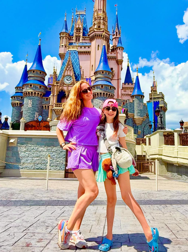 A person and a child stand together smiling in front of Cinderella Castle at Disney World on a bright, sunny day.