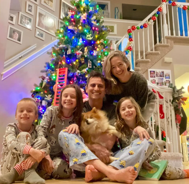 A family of five and their dog sit in festive pajamas on the floor in front of a colorful, lit-up Christmas tree.