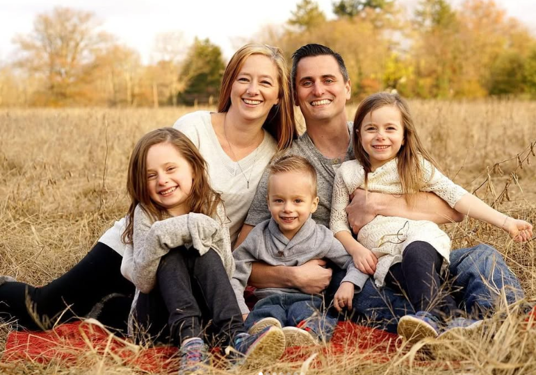 A family of five sitting together on a red blanket in a grassy field during autumn, smiling at the camera.