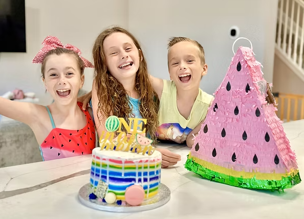 Three happy children posing behind a colorful striped cake and a large watermelon-shaped piñata.