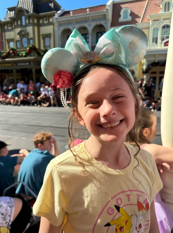 A smiling child wearing teal mouse ears with a flower and pearl accessory, in front of a Disney park building.