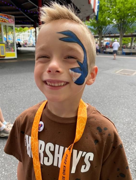 A smiling child wearing a Hershey’s shirt with blue shark face paint on one side, standing in an outdoor park.