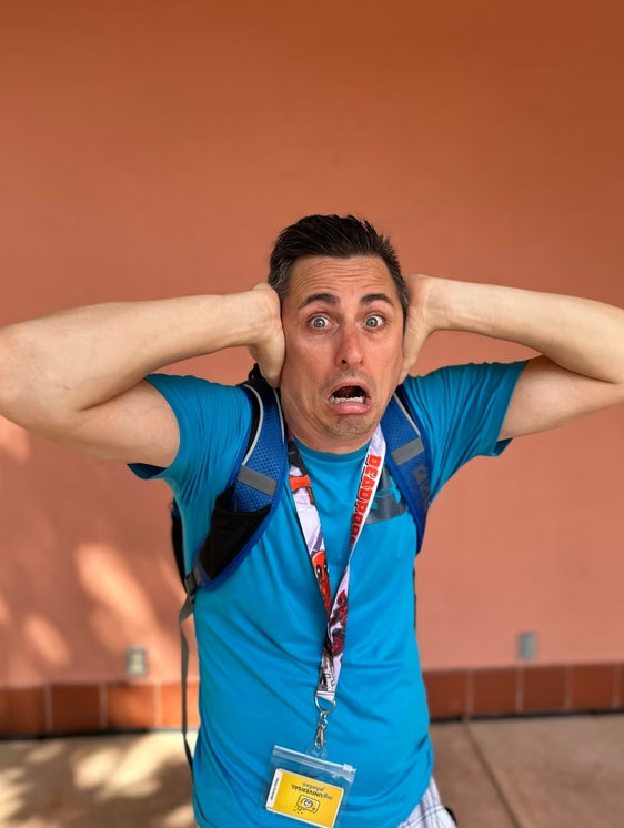 A person in a blue shirt with a backpack and lanyard holds their hands over their ears with a wide-eyed, shocked expression.