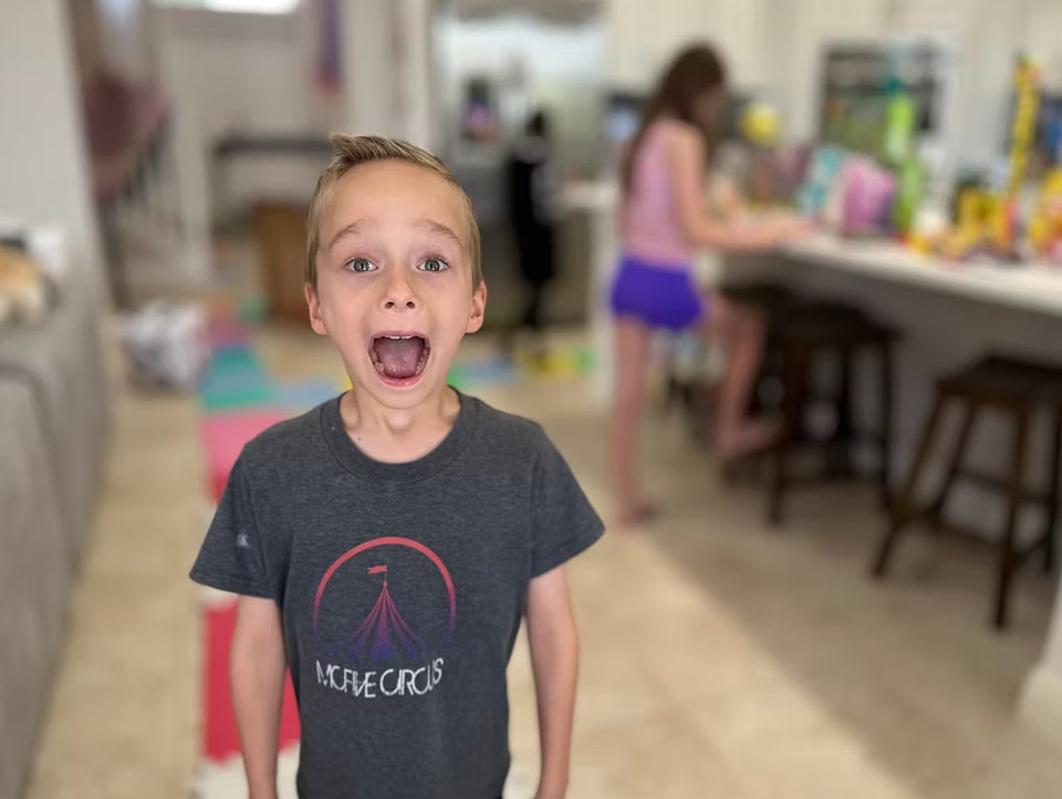 A young person with an excited expression stands in the foreground, while another person sits at a kitchen counter behind.