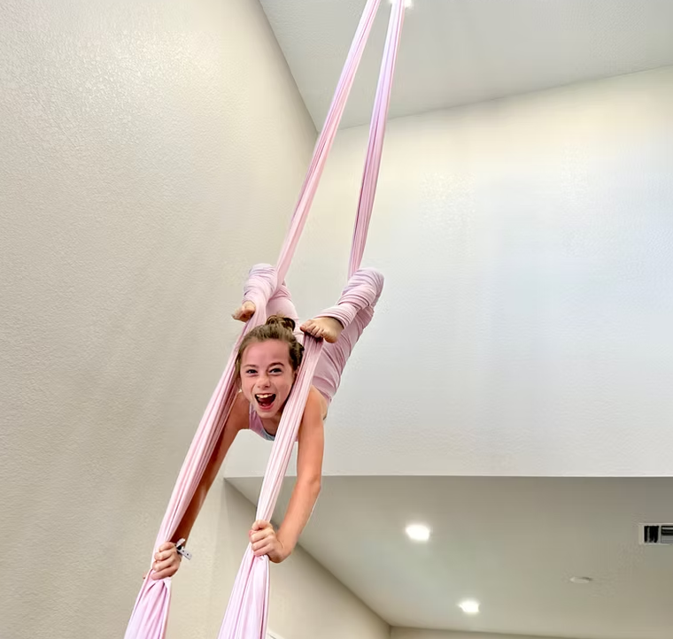 A person wearing light pink clothing hangs upside down while performing a pose on pink aerial silks in a bright room.