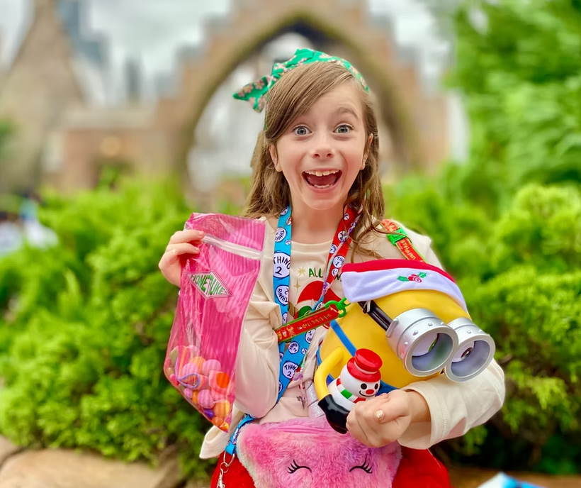 A smiling child holding a pink bag of treats and a Minion toy in front of a stone archway at a theme park.