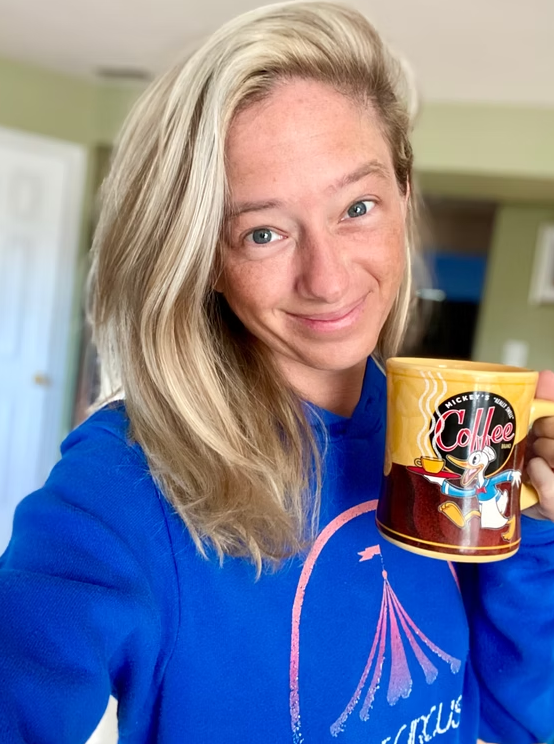 A person with blonde hair wearing a bright blue hoodie smiles while holding a yellow mug featuring Donald Duck.