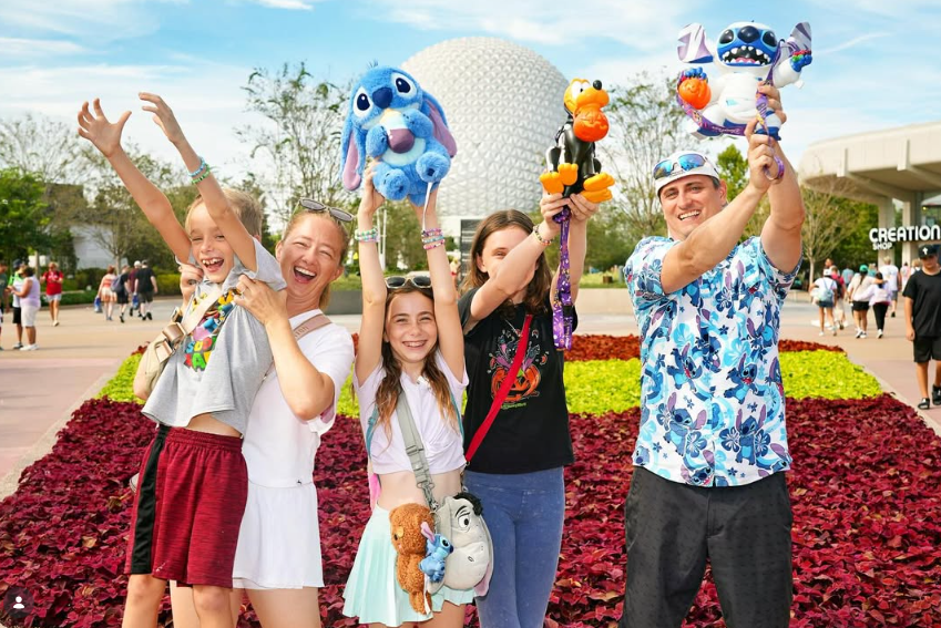A family holds up plush toys in front of the Epcot sphere at Disney World, smiling and laughing under a sunny sky.