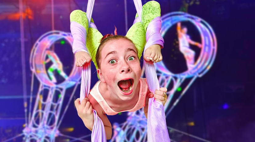 An aerialist with an excited expression hangs from lavender silks in front of blurred, glowing spinning wheels.