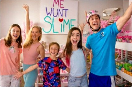 Smiling group standing under a colorful “ME WONT SLIME” sign, with raised hands in a bright room.
