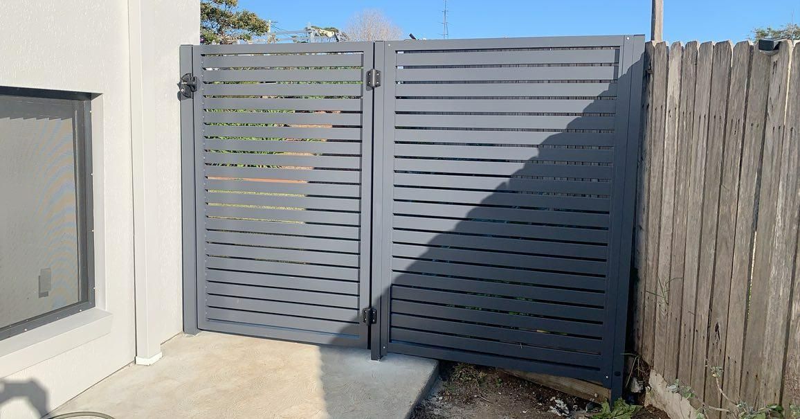 A Gray Gate Is Sitting Next to A Wooden Fence — Optimum Aluminium Fabrication in Dapto, NSW