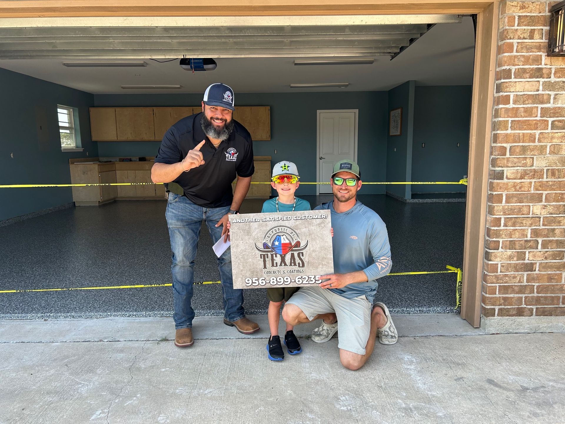 Three people in a garage holding a sign. Two adults, one child; shiny floor, sign has state logo.