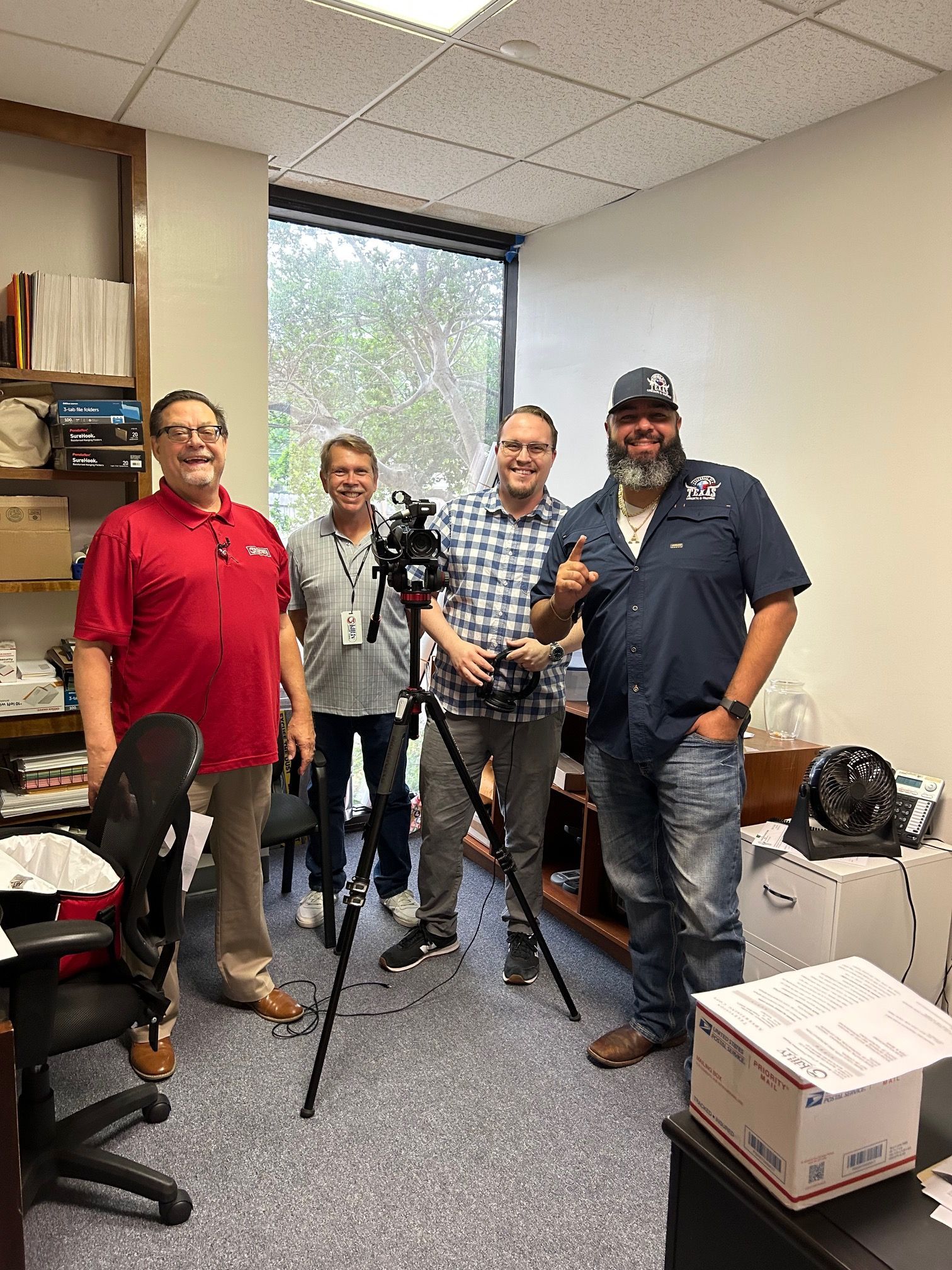 Four men in an office setting, one holding a camera on a tripod, smiling.