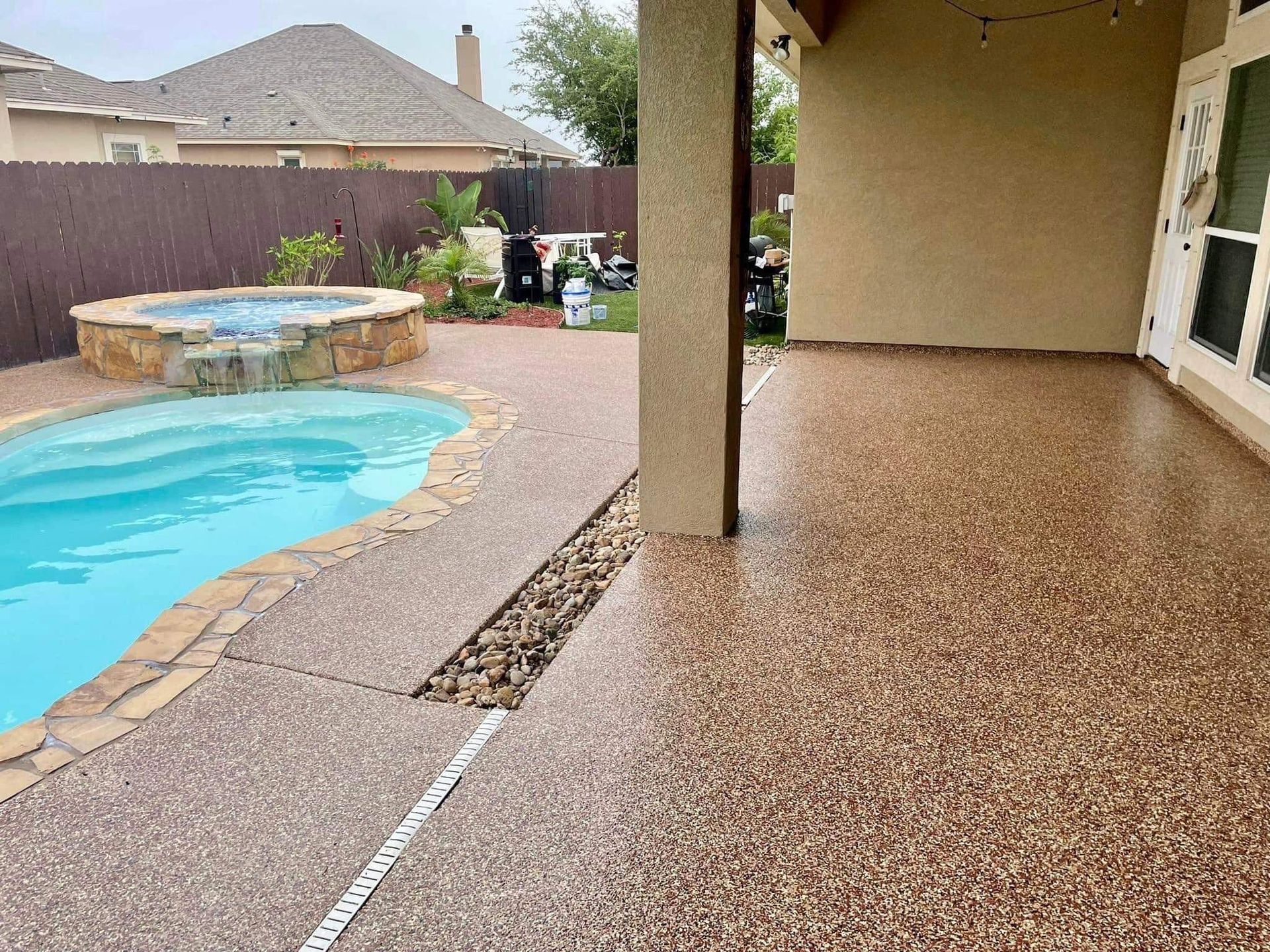Backyard with a pool, jacuzzi, and textured concrete patio; brown and tan colors dominate.