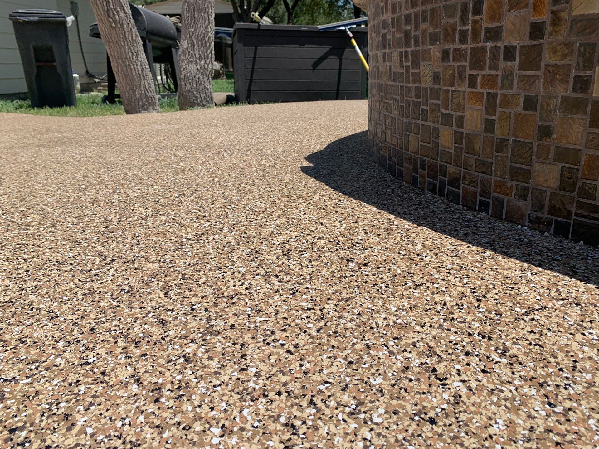 Gravel-like surface with tan, black, and white chips next to a mosaic tile wall and trees.