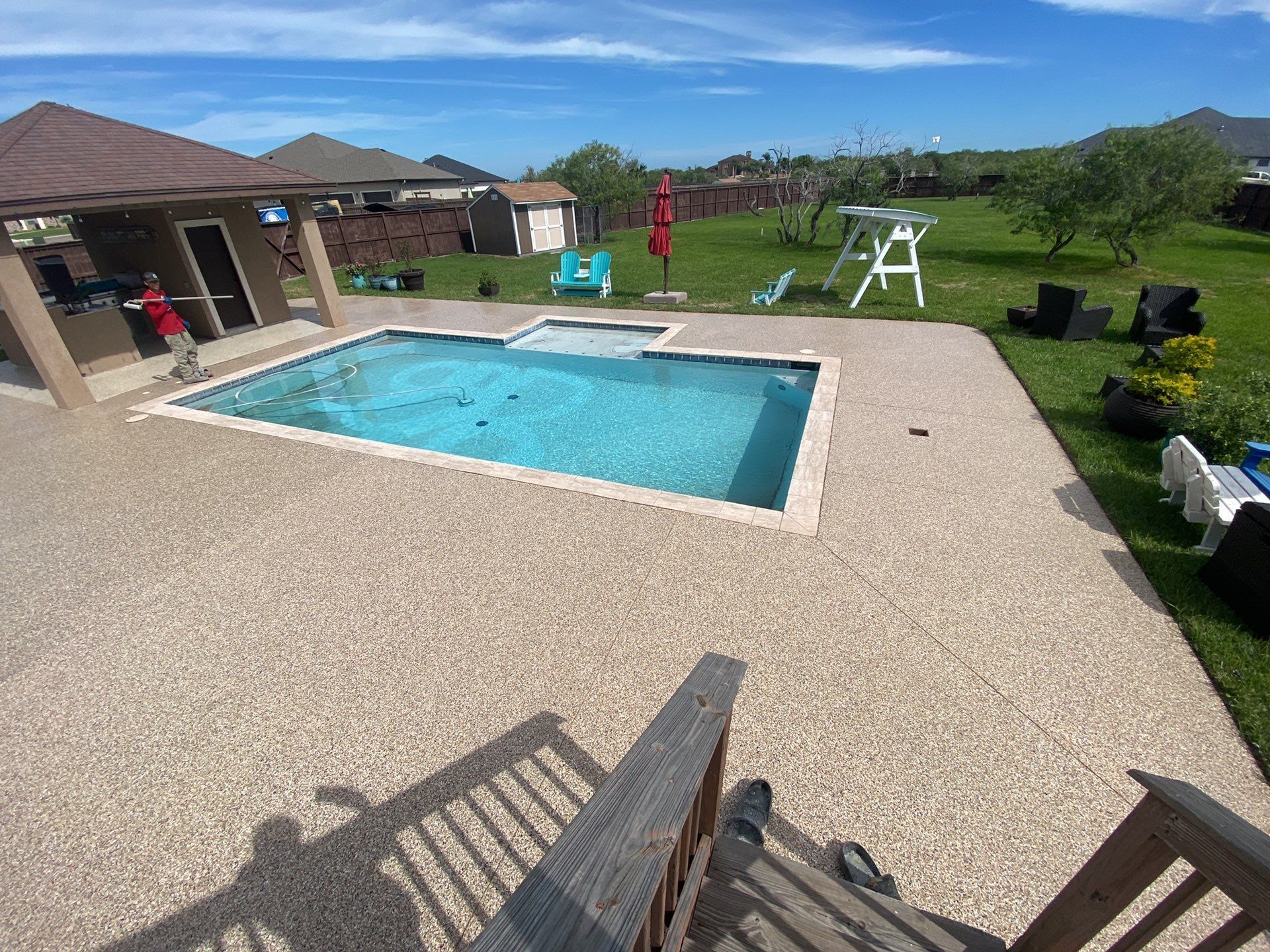 Pool area with beige concrete, a blue pool, and a brown gazebo under a blue sky.