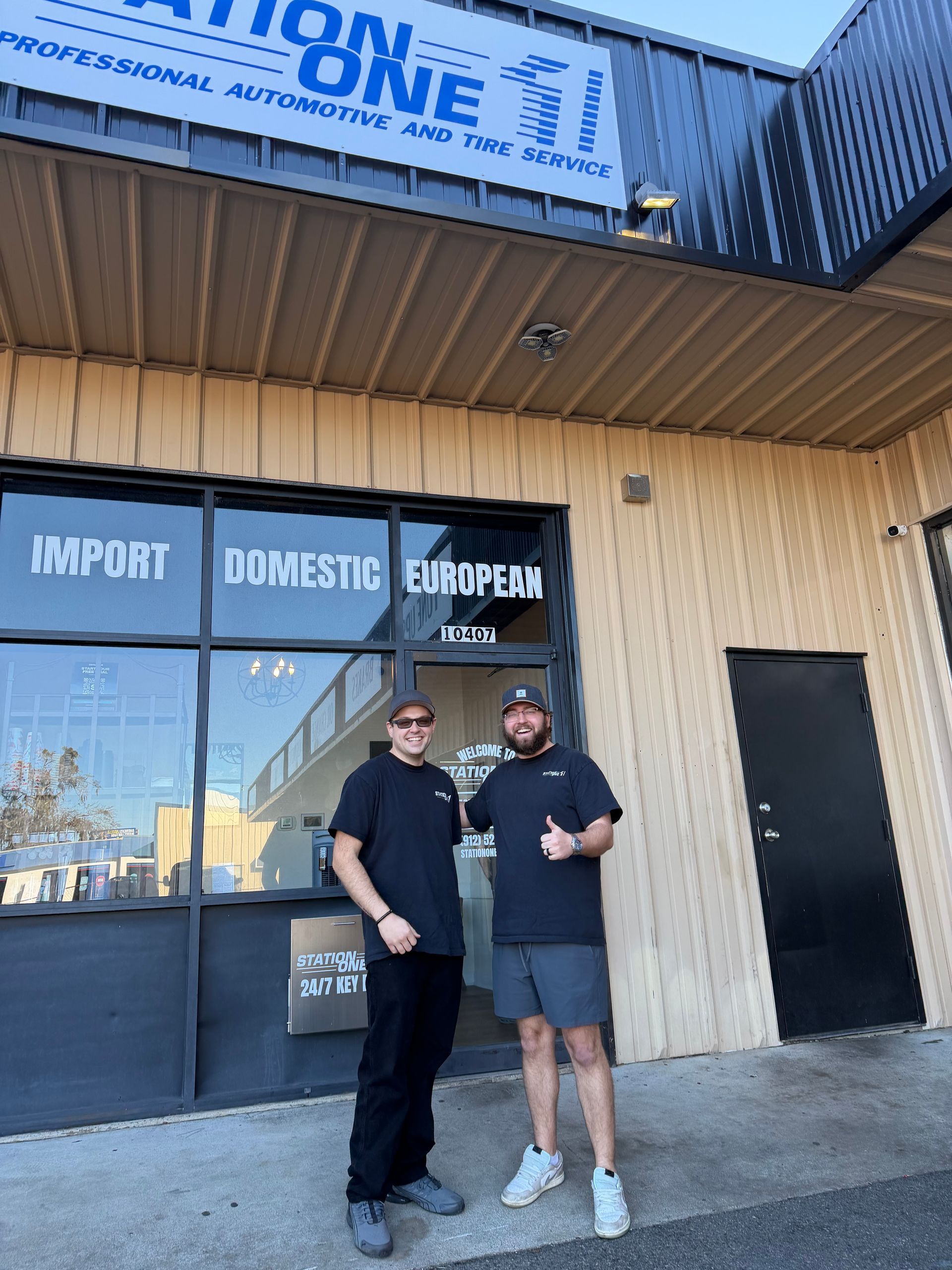 Two men standing outside Station One, a professional automotive shop, with the man on the right giving a thumbs up.