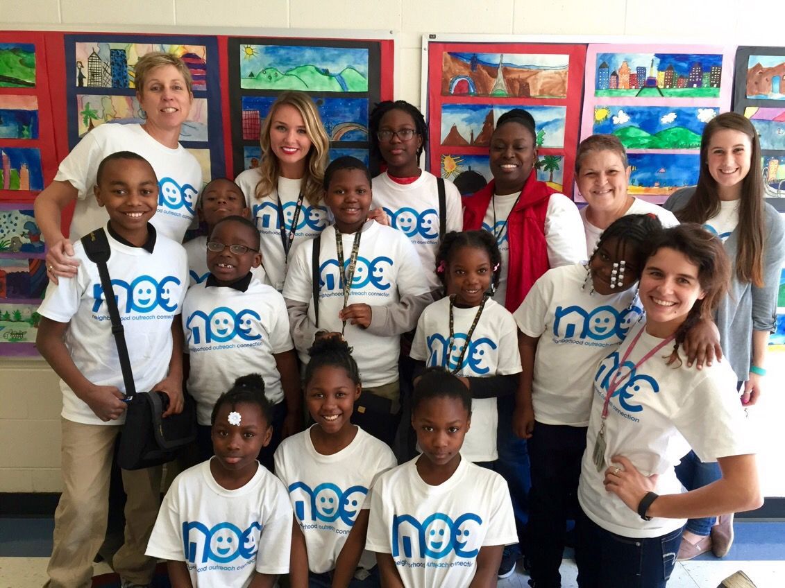 A diverse group of people wearing matching white t-shirts with a blue smiley logo pose together in an art-filled classroom.