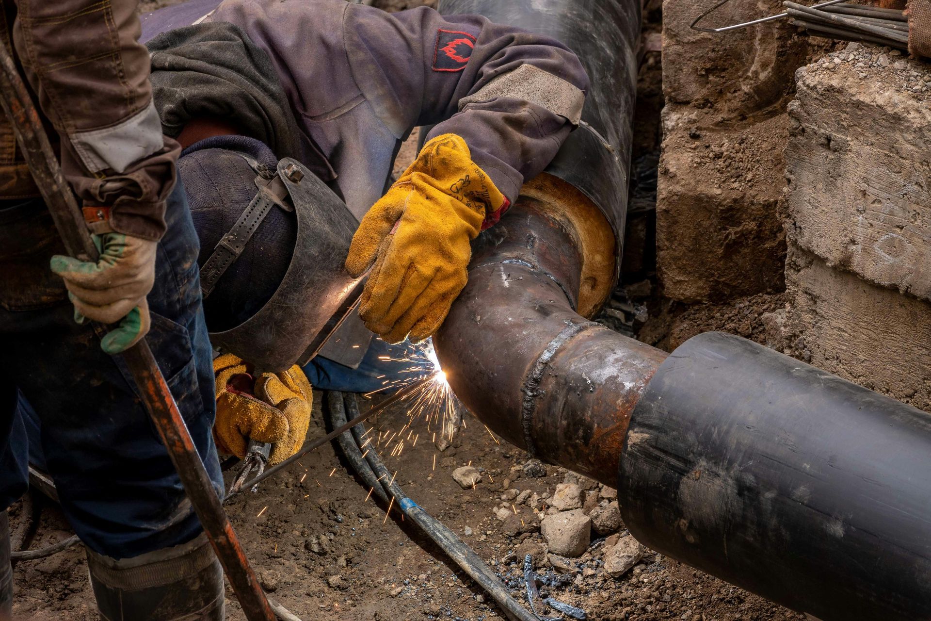 Welder in yellow gloves welding a metal pipe elbow in a construction setting, sparks flying.